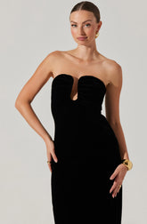 A woman is wearing a strapless, form-fitting black dress with a large keyhole cutout in the center of the bust. The dress has a sleek, velvet-like texture with subtle horizontal stitching details across the bust area. She accessorizes with gold jewelry, including earrings, a bracelet, and rings.